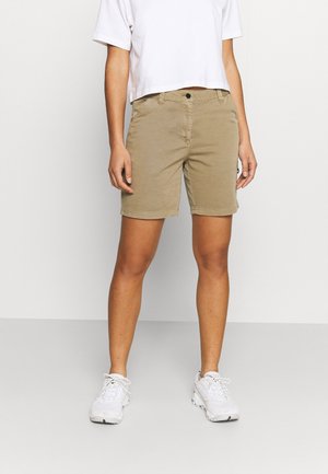 Personne portant un short beige jusqu'aux genoux, un t-shirt court blanc et des baskets blanches, se tenant devant un fond clair uni.