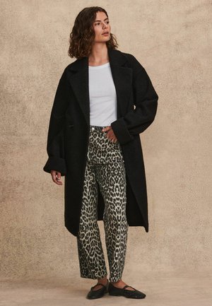Čierny, nadrozmerný vlnený kabát s širokými klopami, nosený cez bielu košeľu a béžové nohavice s leopardím vzorom. K outfitu sa hodia čierne slip-on topánky.