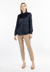 Blusa di raso blu navy con maniche lunghe, chiusura con bottoni e tasca sul petto; abbinata a pantaloni beige a sigaretta e tacchi nudi.
