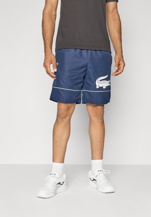 Pantalones cortos deportivos azul marino con acentos blancos y un gran logo de cocodrilo, hechos de un tejido ligero, combinados con zapatillas deportivas blancas y calcetines.