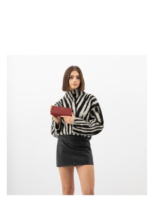 Femme portant un pull à rayures zèbre et une jupe noire, tenant un portefeuille rouge sur un fond blanc uni.