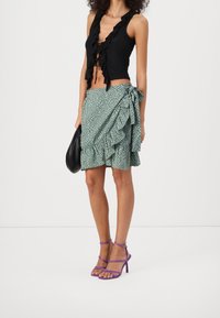 Haut noir sans manches avec bords froncés, associé à une jupe portefeuille verte à motifs avec volants, et des talons hauts à brides violets.