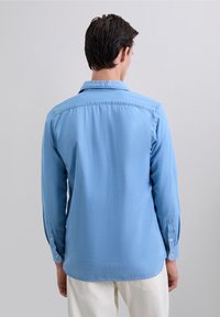 Hombre vestido con una camisa de manga larga azul claro y pantalones blancos, visto desde la espalda sobre un fondo gris plano.