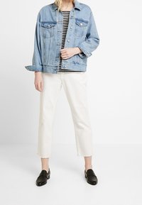 Giacca di jeans, azzurro chiaro, taglio rilassato con due tasche sul petto e chiusura a bottoni. Camicia a righe sotto. Pantaloni bianchi a metà gamba e scarpe nere slip-on.