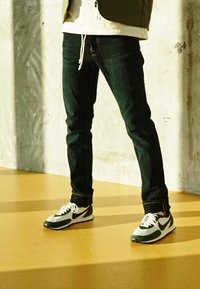 Jeans de ganga escuros com um corte justo, combinados com ténis Nike verdes e brancos com detalhes pretos e atacadores brancos sobre um chão claro.