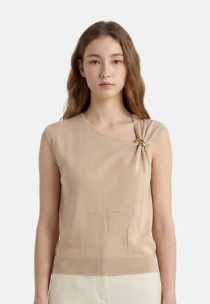 Giovane donna con lunghi capelli castani indossa una canotta beige con un dettaglio arricciato tenuto da un anello dorato sulla spalla sinistra.