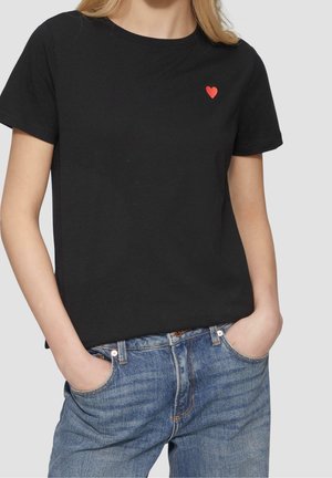 Osoba ubrana w czarną koszulkę z krótkim rękawem z małym czerwonym sercem na lewej piersi, w połączeniu z niebieskimi dżinsami, ręce w kieszeniach.