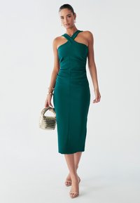 Vestido midi ajustado verde con tirantes cruzados, tela texturizada y detalles de fruncidos. Combinado con un pequeño bolso tejido y sandalias nude.