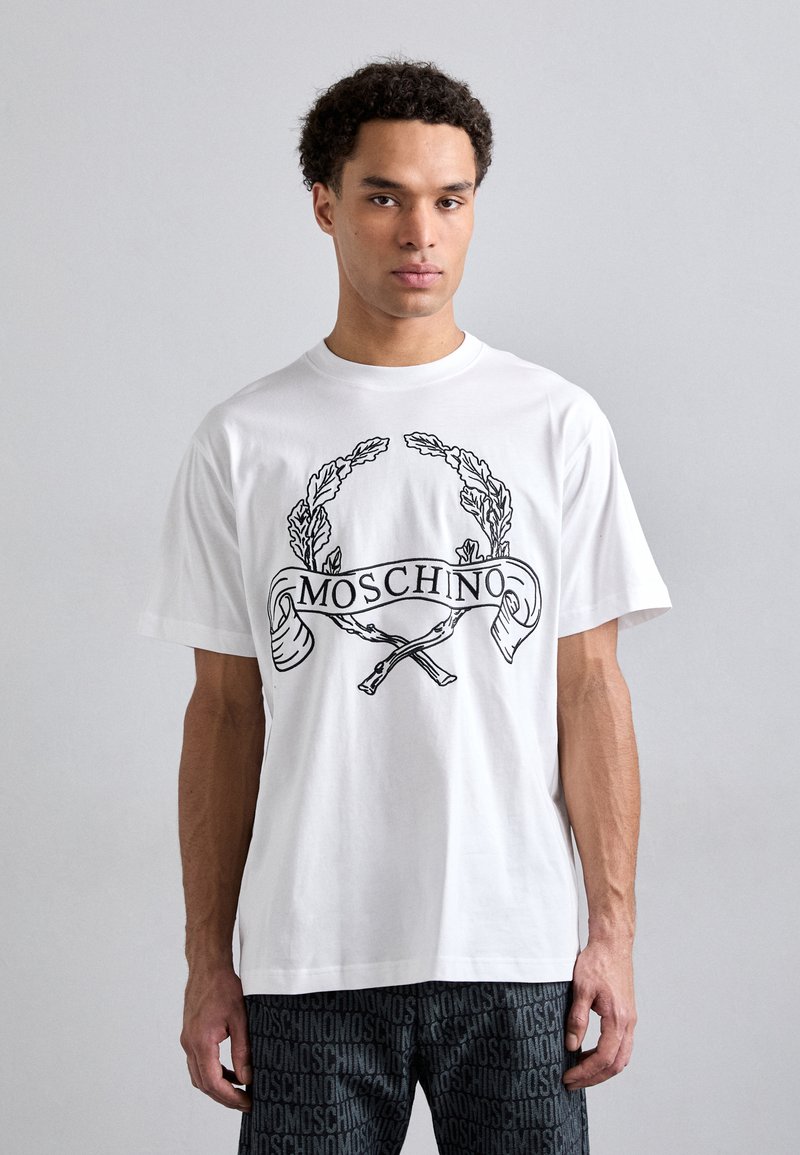 T-shirt en coton blanc avec un design graphique noir mettant en avant des feuilles de laurier et le texte "MOSCHINO" sur la poitrine. Coupe ample.
