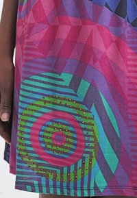 Main à la peau foncée à côté d'une robe colorée avec des motifs géométriques roses, violets, verts et bleus, ornée de petites perles rouges sur un motif circulaire.