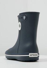 Botte de pluie noire mi-mollet en Crocs avec une bordure blanche, détail de trous circulaires près du haut, et étiquette "crocs" au niveau du talon.