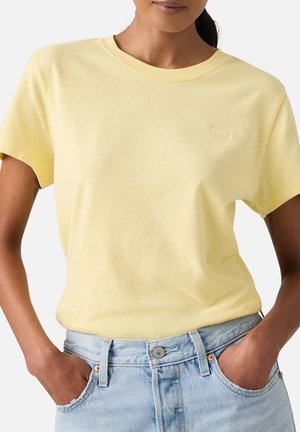 Femme portant un t-shirt jaune pâle à manches courtes rentré dans un jean en denim bleu clair, mains dans les poches, debout devant un fond uni.