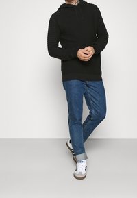 Man med svart ribbad huvtröja, blå uppvikt jeans och vita Adidas-sneakers med svarta ränder, som står mot en vit bakgrund.