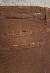 Tejido de mezclilla marrón con una textura suave, que presenta un bolsillo trasero y un detalle de costura estándar. La cinturilla tiene un lazo para cinturón con un diseño de pespuntes.