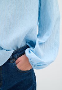 Ljusblå långärmad skjorta med en strukturerad mönster, rynkade ärmslut och avslappnad passform, i kombination med mörka denimjeans.