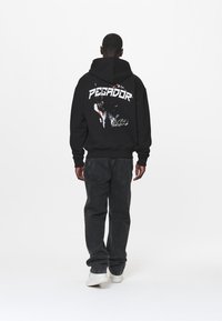 Sudadera negra con capucha que presenta el texto blanco "PEGADOR" y un gráfico de un lobo en la parte posterior. Combinada con jeans oscuros y zapatillas blancas.