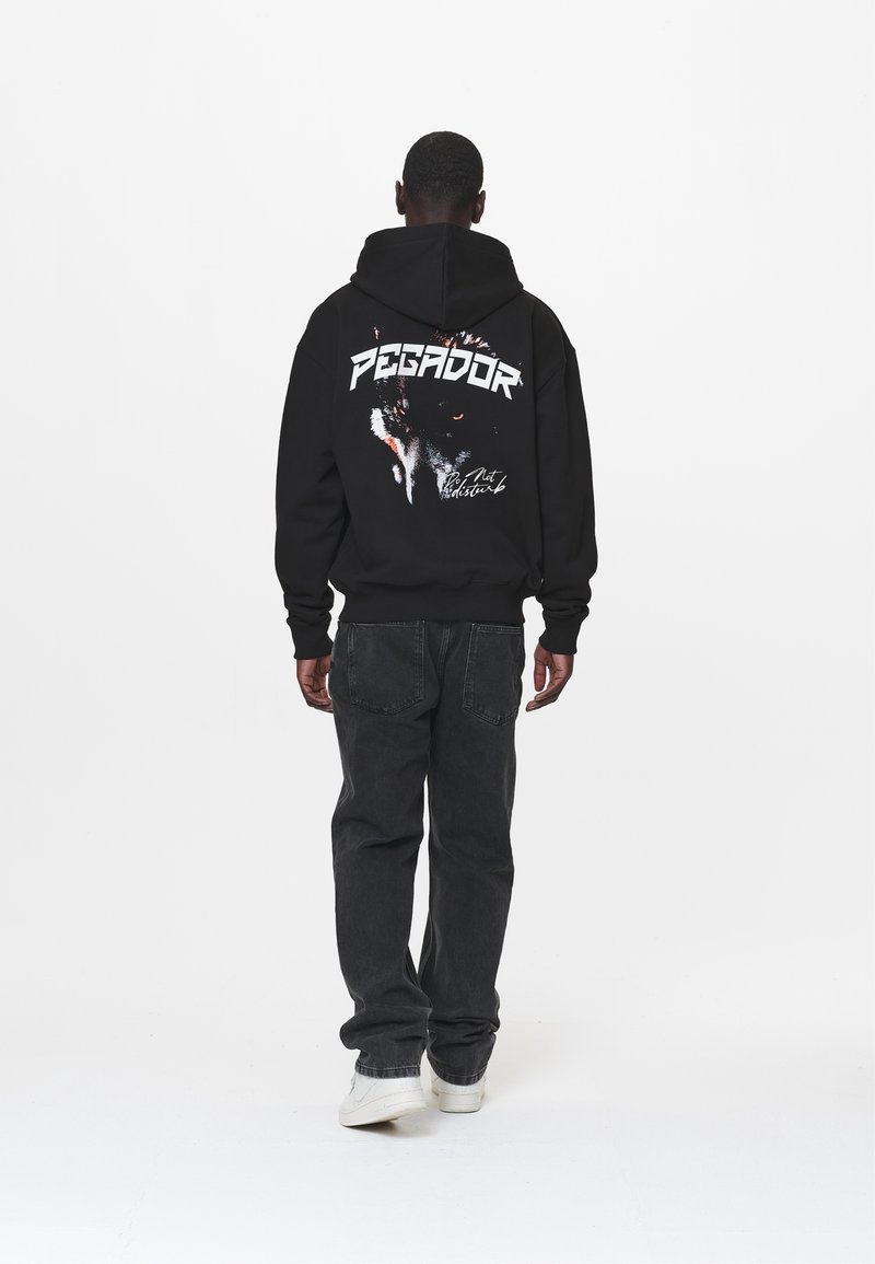 Sudadera negra con capucha que presenta el texto blanco "PEGADOR" y un gráfico de un lobo en la parte posterior. Combinada con jeans oscuros y zapatillas blancas.