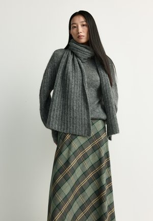 Frau mit langen dunklen Haaren, die einen grauen Strickpullover und Schal trägt, kombiniert mit einem grünen und braunen karierten Rock, stehend vor einem schlichten Hintergrund.