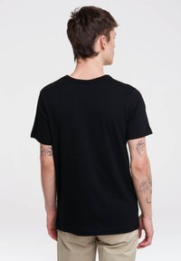 Camiseta negra de manga corta hecha de algodón, con cuello redondo y un corte relajado. Se puede ver un tatuaje en el brazo derecho.