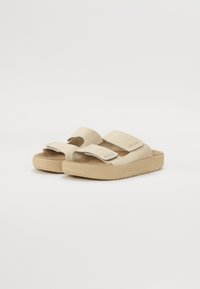 Beiga sandaler med två justerbara remmar, mjukt mockamaterial och texturerade gummisulor med ett vågigt mönster. Logotyp på en rem.