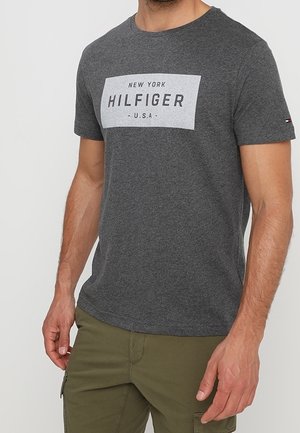 Man wearing dark gray short-sleeve T-shirt with "New York Hilfiger U.S.A" text and olive green cargo pants, standing against plain background.