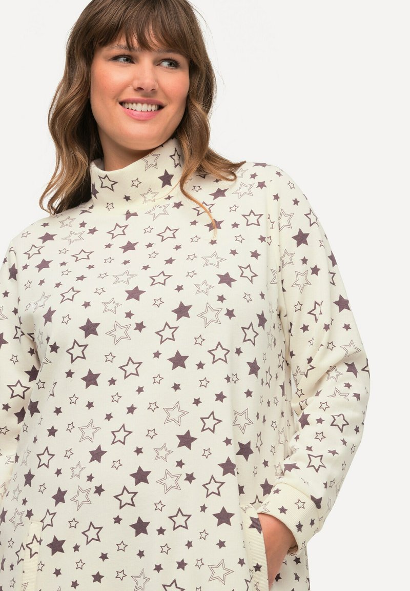 Maglia a maniche lunghe di colore crema con motivo a stelle in marrone scuro. Collo alto, vestibilità comoda, polsini a costine e tasche laterali ne migliorano la funzionalità.