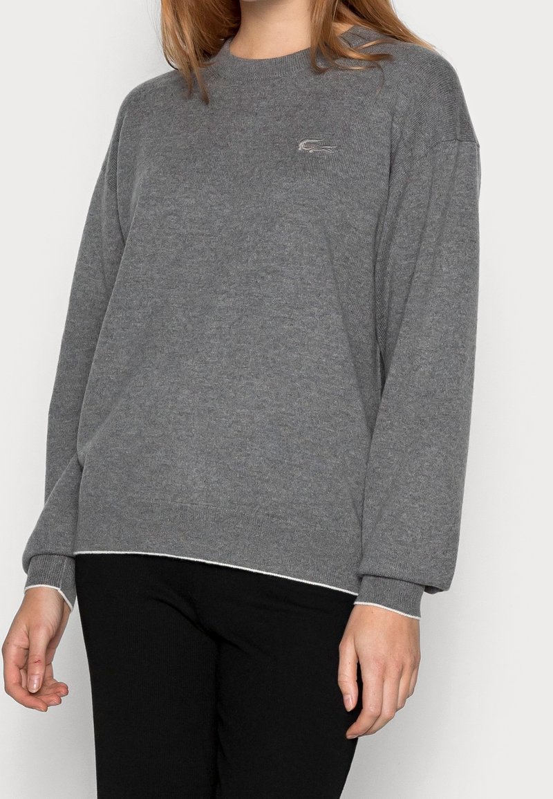 Femme portant un pull gris à manches longues avec un petit logo crocodile sur la poitrine et un pantalon noir, debout devant un fond clair.