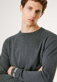 Suéter gris de punto con un cuello redondo y mangas largas. Presenta puños acanalados que exhiben un nombre de marca en texto blanco.