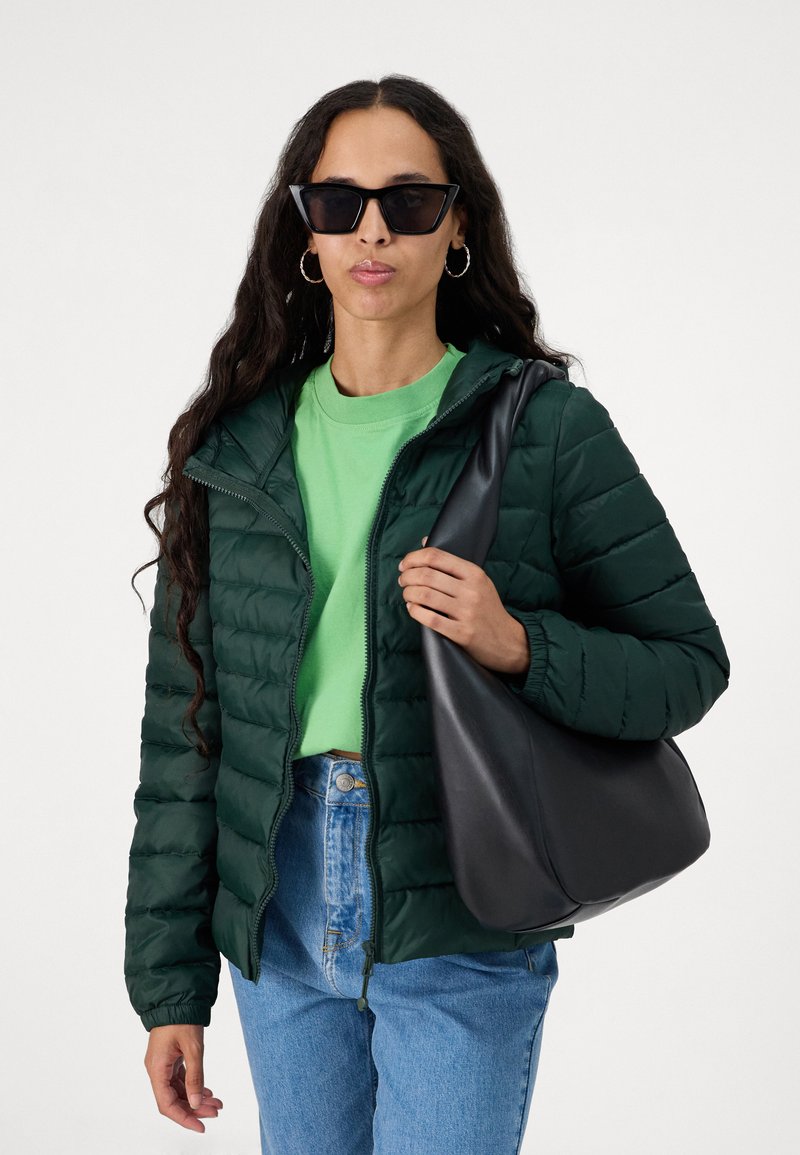 Groene gewatteerde jas met rits, lange mouwen en elastische manchetten; gecombineerd met een groen T-shirt en high-waisted blauwe jeans; houdt een zwarte tas vast.