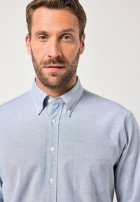 Camisa de botón azul claro en una tela texturada, con cuello puntiagudo, bolsillo frontal y botones blancos.