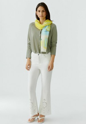 Woman wearing olive green cardigan, white pants with cutout patterns, white heeled sandals, and yellow-blue scarf standing against a plain background.