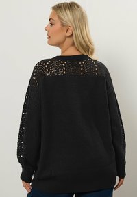 Svart tröja med ribbat mönster och öppen spetsdetaljering på ärmarna och övre ryggen, med en lös, avslappnad passform och rund halsringning.