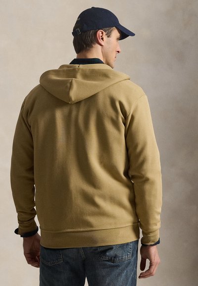 Homme portant un sweat à capuche beige, un jean bleu et une casquette bleu foncé, de dos sur un fond neutre.