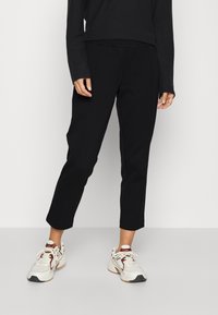 Sorte tapered bukser med et glat stof, der har en mellemhøj talje og diskrete forkertninger. Kombineret med hvide og brune sneakers.
