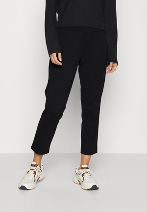 Pantalons noirs fuselés en tissu lisse, dotés d'une taille mi-haute et de plis subtils à l'avant. Associés à des baskets blanches et brunes.