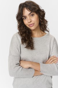 Svetlo siva jopica z žepi s rebrastimi manšetami, mehko teksturo in sproščenim krojem. Model stoji z arms crossed, s čimer prikazuje detajle izreza in rokavov.
