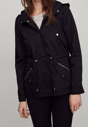 Femme portant une veste à capuche noire avec une fermeture éclair sur le devant, des cordons à la taille et des poches zippées par-dessus un haut gris clair.