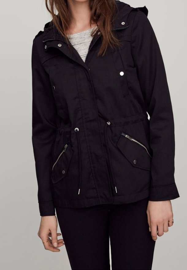 Femme portant une veste à capuche noire avec une fermeture éclair sur le devant, des cordons à la taille et des poches zippées par-dessus un haut gris clair.