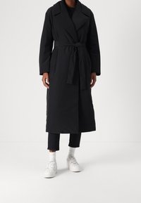 Black long coat with a belt, tailored design, wide collar, and side pockets, worn over dark pants and white sneakers with textured fabric.