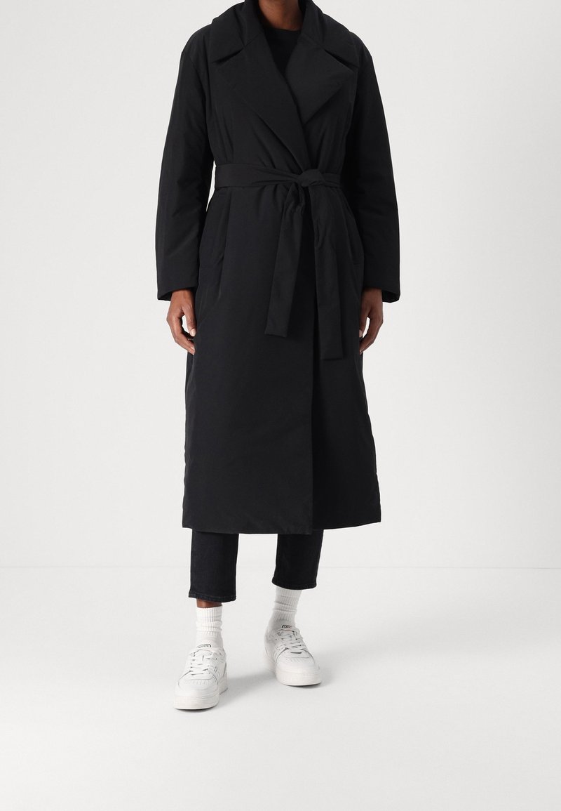 Black long coat with a belt, tailored design, wide collar, and side pockets, worn over dark pants and white sneakers with textured fabric.