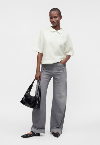 Femme debout, portant un haut blanc à manches courtes, un jean gris à jambes larges, des chaussures noires pointues avec des clous et tenant un sac à main noir brillant.