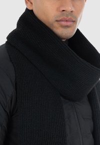 Black knitted scarf with a ribbed texture, wrapped around the neck, complementing a black jacket. Smooth, thick fabric.