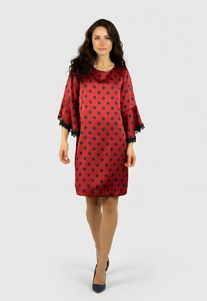 Mujer con cabello largo y rizado, vestida con un vestido rojo hasta la rodilla con lunares negros y encaje negro en las mangas anchas, caminando hacia adelante.