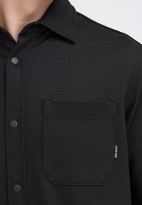 Primer plano de una persona que lleva puesta una camisa negra texturizada abotonada, con cuello y un bolsillo en el pecho etiquetado como "BLEND".