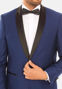 Giacca da smoking blu strutturata con rever in satin nero, camicia bianca e papillon; chiusura a un bottone e tasche laterali visibili.