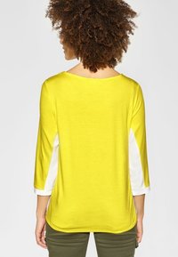 Gul topp med trekvartslång ärm och vita sidopaneler, slät textur, avslappnad passform och rund halsringning, sedd bakifrån.