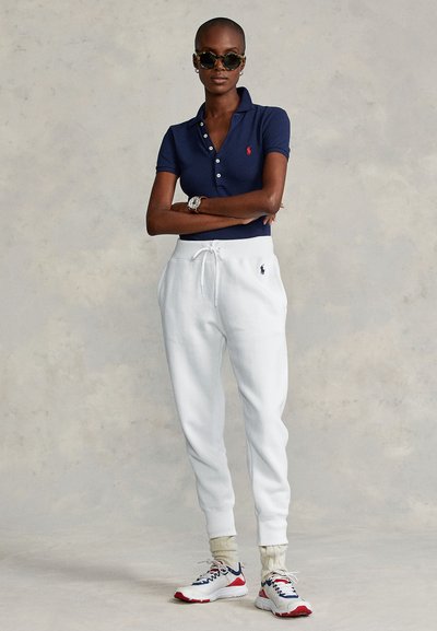 Γυναίκα με μπλε σκούρο πόλο μπλουζάκι, λευκές φόρμες με κορδόνι, λευκές κάλτσες, πολύχρωμα αθλητικά παπούτσια και γυαλιά ηλίου, στέκεται με τα χέρια σταυρωμένα.