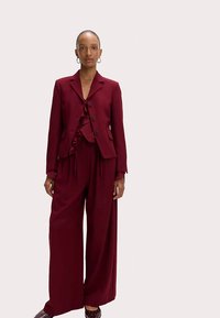 Bordeauxkleurig op maat gemaakt pak met een gerafelde top, bestaande uit een getailleerde blazer en wijde broekspijpen, gemaakt van gladde stof. Model staat rechtop.