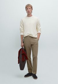 Heller Cremefarbener Pullover, khakifarbene Hose und eine dunkle Tasche mit einem roten Kleidungsstück. Die Materialien wirken weich, mit minimalen Details und einer lockeren Passform.