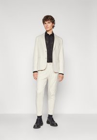 Lys beige skreddersydd dress med en ensfarget blazer og matchende slim-fit bukser, kombinert med en svart mønstret skjorte og svarte sko.